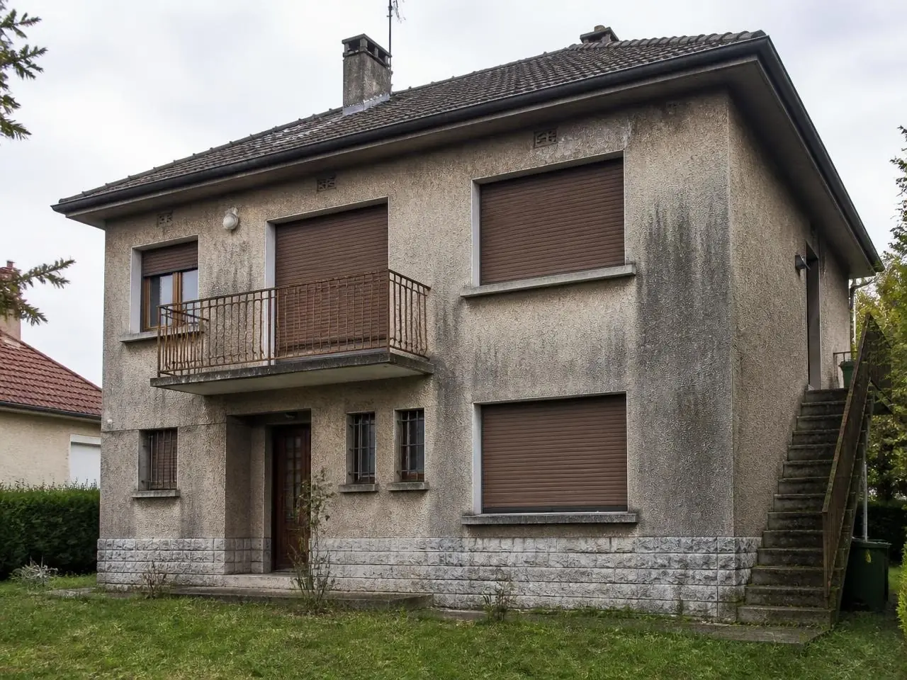 Maison à étage au crépi grisé et marqué de coulures, volets roulants bruns, soubassement en pierre